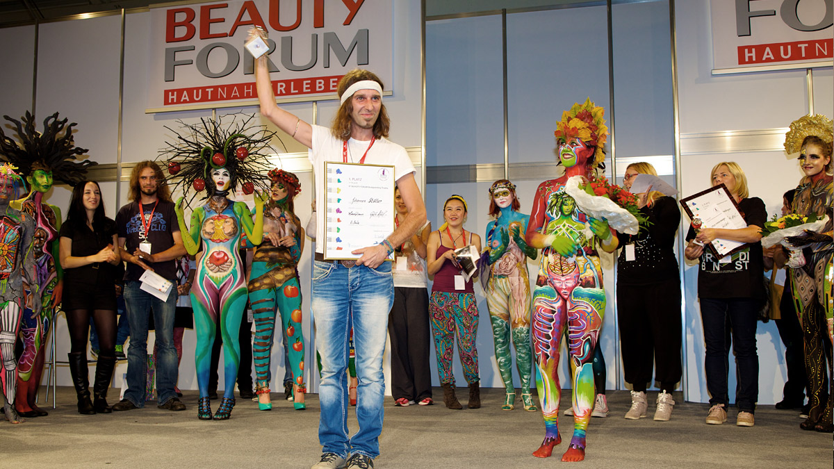 Siegerehrung beim Beauty Forum in München: Ein Künstler hält seine Urkunde und einen Preis in die Höhe, während bemalte Models und weitere Teilnehmer auf der Bühne stehen.