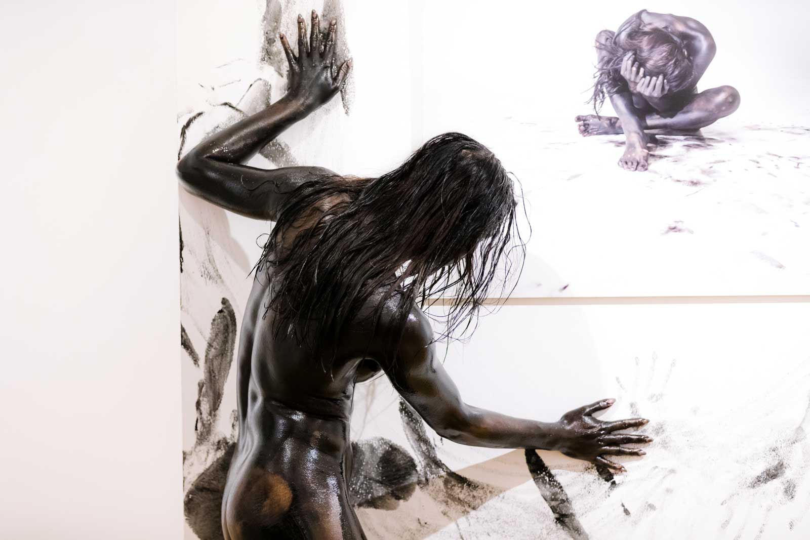 Körperbemalte Person mit schwarzer Farbe lehnt mit Händen und Körper an eine weiße Wand, die mit Handabdrücken bedeckt ist. Im Hintergrund hängt ein Foto einer ähnlichen Figur in zusammengesunkener Pose.
