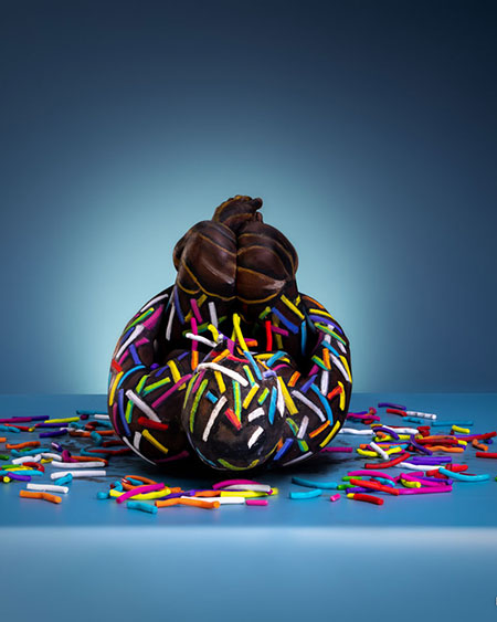Menschliche Figur, kunstvoll bemalt und posiert wie ein Schokoladendonut mit bunten Zuckerstreuseln, vor blauem Hintergrund.