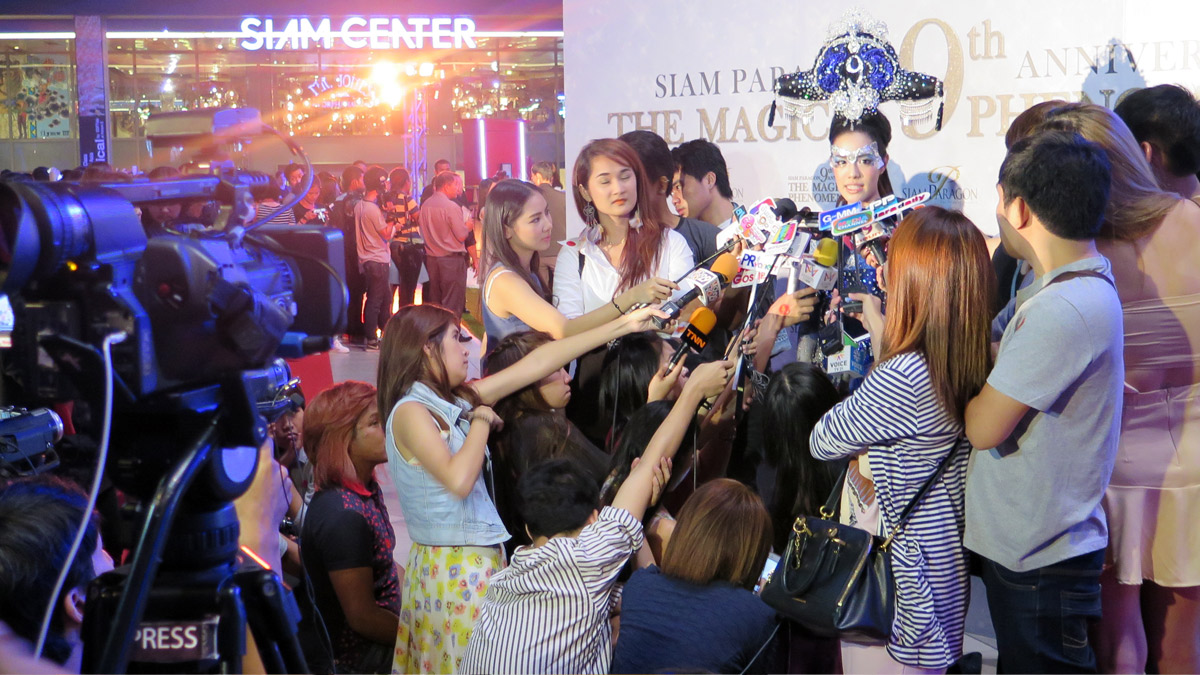 Presseinterview bei einer Veranstaltung im Siam Center in Thailand: Eine Frau mit auffälligem Kopfschmuck und Bodypainting wird von zahlreichen Reportern und Kameras umringt, während viele Mikrofone auf sie gerichtet sind.