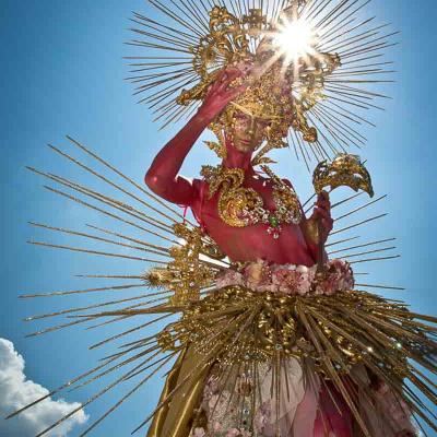 Performer with red-painted skin wearing an elaborate gold headdress and spiked halo costume, standing outdoors against a bright blue sky.