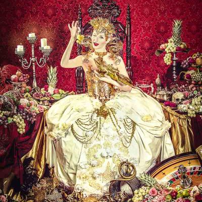 Performer dressed as a lavish queen in a gold gown and crown, seated on a throne amid fruit, candles, skulls, and scattered treasures against red patterned wallpaper.