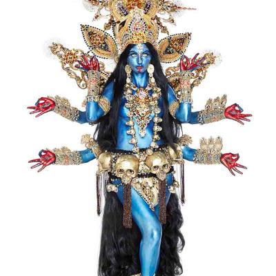 Performer painted blue as a multi-armed goddess, wearing an ornate gold headdress, skull belt, and elaborate jewelry, standing against a white background.