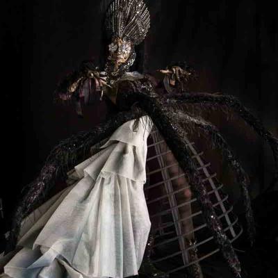 Performer in a dramatic black and white gown with a cage-style skirt, wearing an ornate tall headpiece and long dark textured sleeves, posed against a dark background.