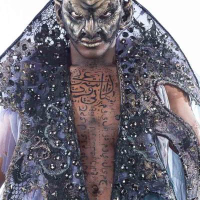 Performer in an elaborate dark costume with horned mask, ornate cape, and script painted across the chest, staring intensely at the camera.