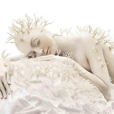 Person painted white with coral-like branches attached to the body, resting on white fabric against a light background.