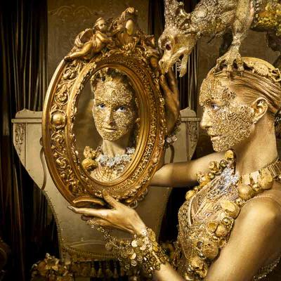 Person in gold body paint and ornate costume holding a gilded mirror, with a dragon sculpture above and candles in the background.