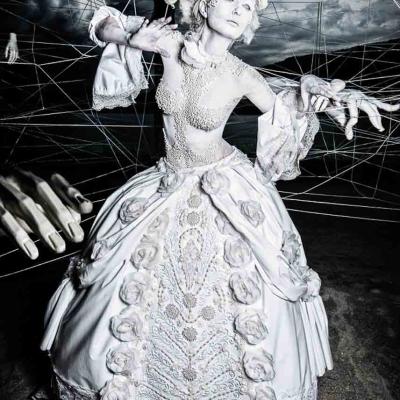 Performer in an ornate white historical-style dress posed like a marionette, surrounded by visible strings and disembodied mannequin hands against a dramatic sky backdrop.