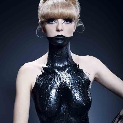 Woman with blonde updo and heavy eye makeup, her neck and torso covered in glossy black paint, against a dark background.