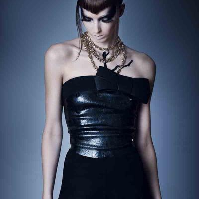 Woman in a black strapless outfit with a glossy bodice and layered gold chains, looking down against a plain dark background.