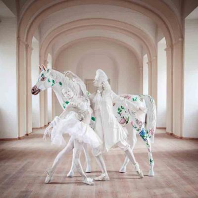 Performer in a flowing white costume posed beside a white horse decorated with painted flowers, inside a large arched hall.