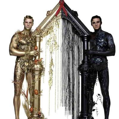 Two men posed as living statues: one painted gold with vines and fruit, the other painted black with dark foliage, each holding a column that forms a peaked frame between them.