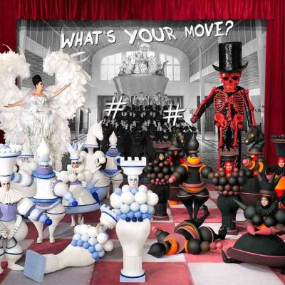 Group of performers in oversized chess piece costumes on a stage with a “What’s Your Move?” backdrop.