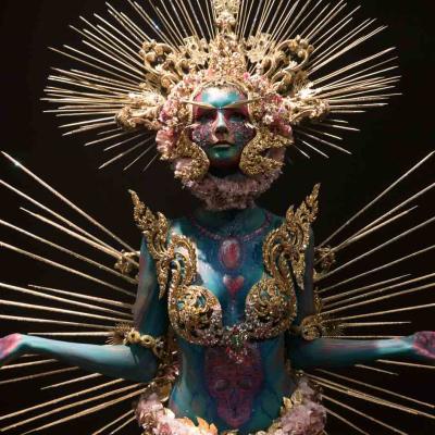 Person with blue-painted body wearing an ornate gold headdress and radiating spikes against a dark background.