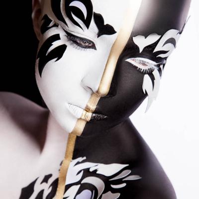 Close-up of a model with split black and white face paint, bold graphic patterns, and a vertical gold stripe down the center.