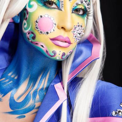 Close-up of a model with colorful fantasy face paint, rhinestone decorations, long white hair, and a blue and purple costume.