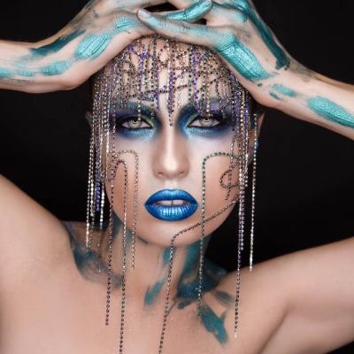 Portrait of a model with blue metallic makeup, crystal chains draped over the face, and hands painted turquoise resting on the head.