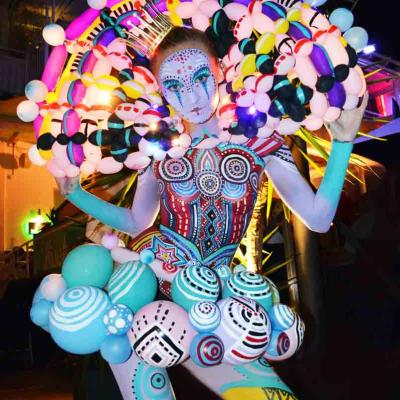 Performer with colorful body paint and sculptural balloon costume, posing at night with bright lights behind.