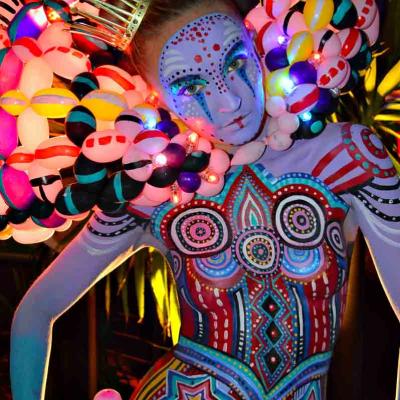Close-up of performer with detailed geometric body paint, wearing a small crown and a glowing balloon headdress.