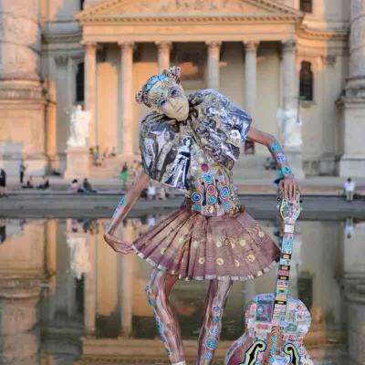 Performer in painted costume posing with a decorated guitar in front of a grand classical building reflected in water.