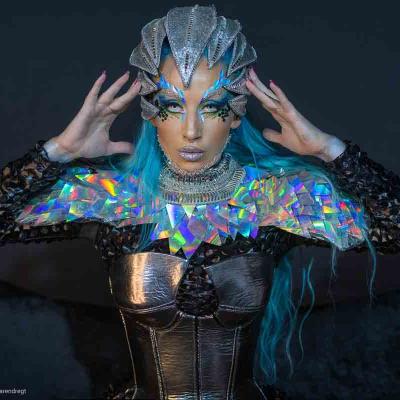 Person with blue hair, metallic headpiece, and holographic collar posing against a dark background.