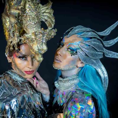 Two people in elaborate metallic fantasy headpieces and dramatic makeup posing against a dark background.