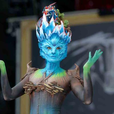 Performer in a blue and green fantasy creature costume with a detailed headpiece posing with raised hands.