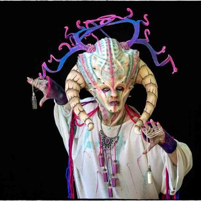 Performer in an elaborate alien-inspired costume with horned headpiece and colorful details posing against a black background.