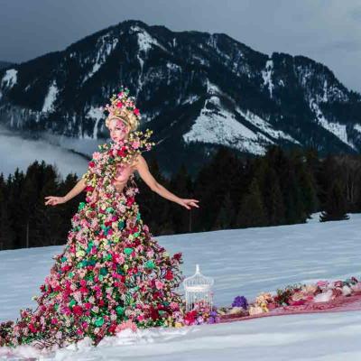 Person in a colorful flower-covered gown standing on snow with mountains in the background.