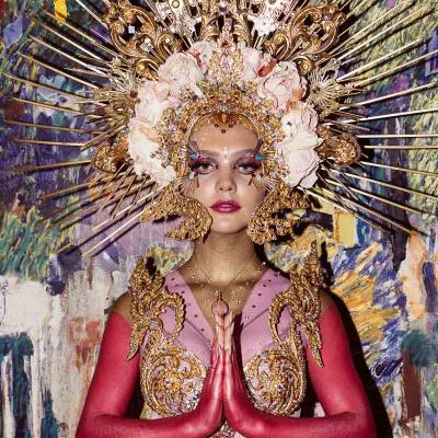 Performer wearing an elaborate gold headdress and ornate costume, posing with hands pressed together in front of a colorful painted background.