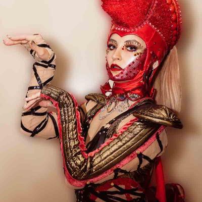 Performer in an elaborate red costume with a glittering horse headpiece posing against a plain background.