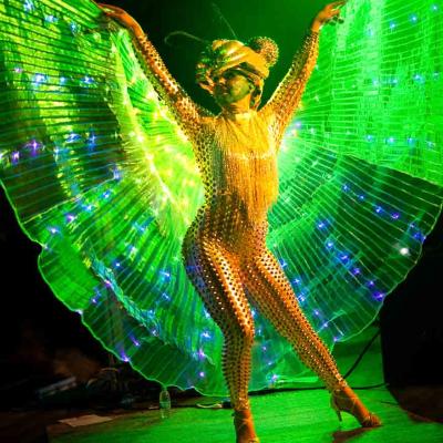 Performer in a gold bodysuit with illuminated green wings dancing on a stage under green lights.