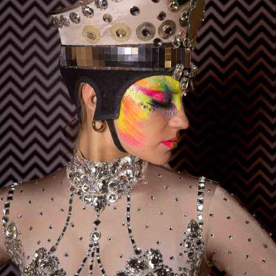Performer in a jeweled costume and metallic headpiece with colorful face paint, posing against a patterned background.