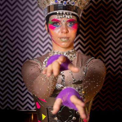 Performer wearing a jeweled headpiece and colorful face paint reaching forward against a patterned background.
