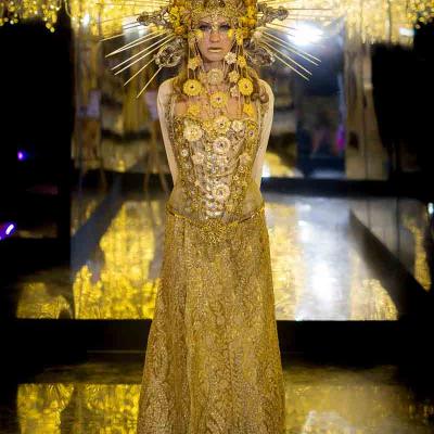 Performer in an ornate gold gown with a radiant headdress standing in a mirrored room with golden lights.