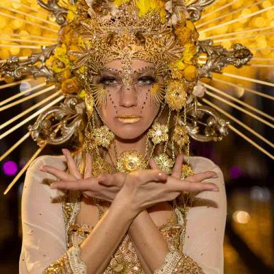 Close-up of a performer in an elaborate gold headdress and jewelry posing with crossed hands against a background of golden lights.