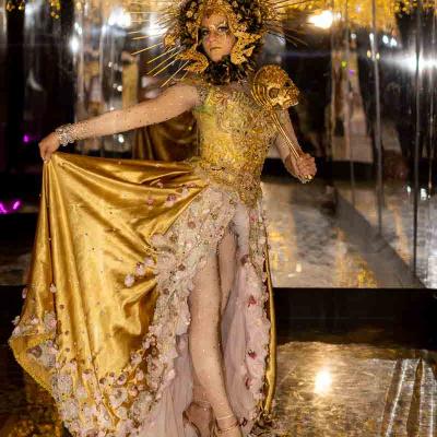 Performer in an elaborate gold costume with a radiant headdress holding a staff, standing in a mirrored room with golden lights.