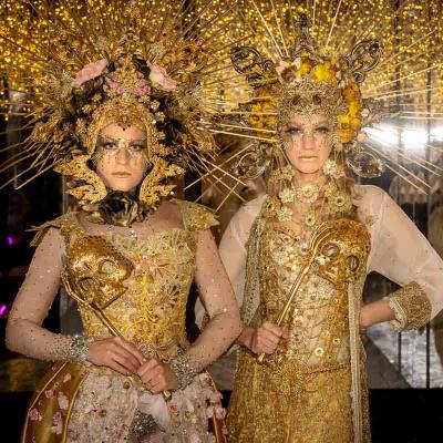 Two performers in elaborate gold costumes with radiant headdresses holding ornate masks in a mirrored room with golden lights.