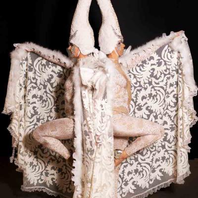 Performer in an elaborate white swan-inspired costume with two long swan neck headpieces and lace wings against a dark background.