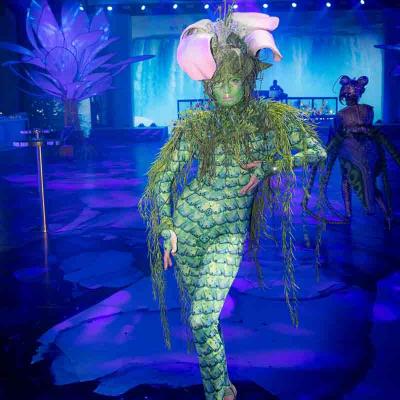 Performer in a green plant-inspired costume with a large flower headpiece walking through a blue-lit event space.