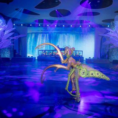Performer in a colorful insect-inspired costume with long curved extensions walking in a blue-lit event space.