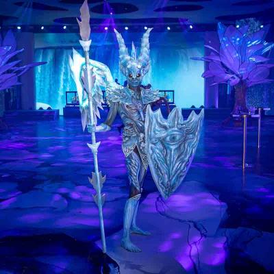 Performer in an icy fantasy armor costume holding a spear and shield, standing in a blue-lit event space.