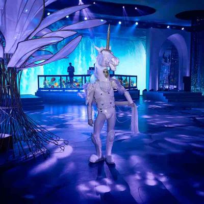 Performer in a white unicorn costume standing in a blue-lit event space with a DJ booth in the background.