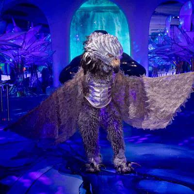 Performer in a large bird-inspired costume with wings spread, standing in a blue-lit event space.