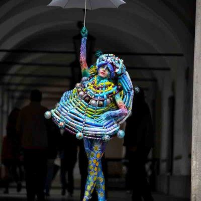 Performer in colorful body paint and a sculptural balloon costume, standing on pointe under a white umbrella in an arched passageway.