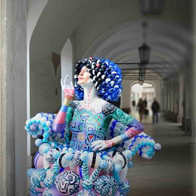 Performer in a colorful, sculptural costume with painted body and curly blue headpiece, posing in an arched hallway.