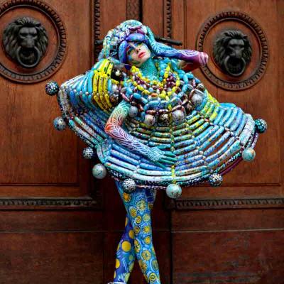 Performer in a colorful, bead-covered sculptural costume and body paint, posing on pointe in front of large wooden doors with lion head carvings.
