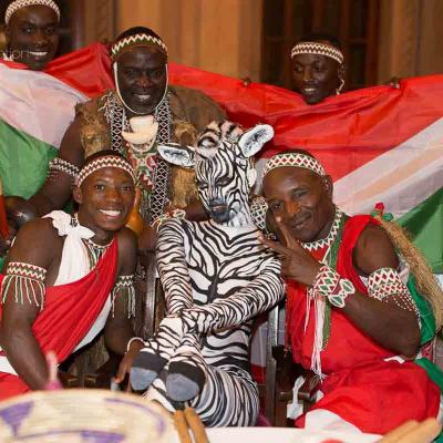 Group of performers in red, white, and green outfits posing with a person painted like a zebra in front of a large red, white, and green flag.