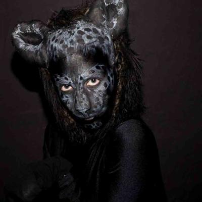 Portrait of a person with dark animal-inspired face paint and rounded ears, posing against a dark background.