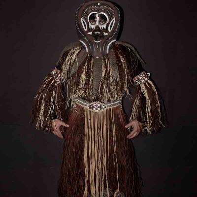 Performer wearing a tall traditional-style mask and a long straw costume standing against a dark background.